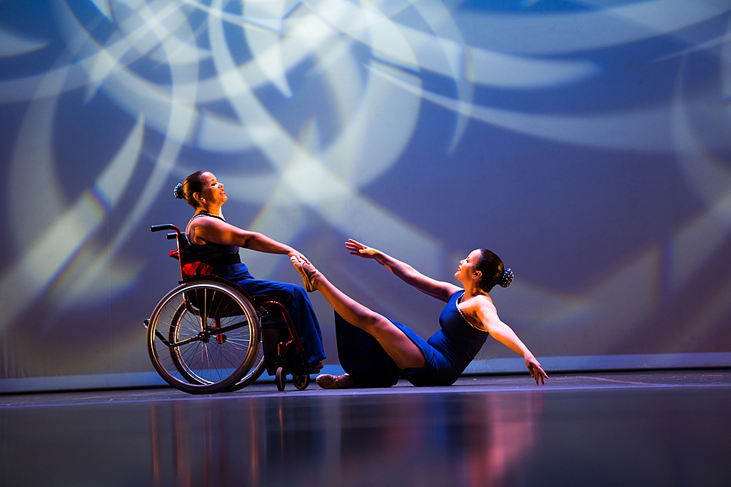 FOTO no palco de duas bailarinas, a esquerda uma cadeirante segura o pé da bailarina da direita que está sentada ao chão de frente para a cadeira com os braços abertos elas se entreolham, a ilumicacão do palco é azul, ao fundo uma projeção de imagens abstratas de raios brancos .