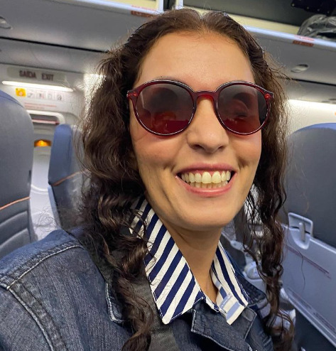 Viviane Garcia, em foto com lindo sorriso, tem cabelos castanhos, usa óculos escuros. Veste uma camisa branca com listas azuis e jaqueta jeans por cima.